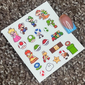 Op de afbeelding: Vel met nagelkunststickers met verschillende personages en elementen uit de Super Mario-videogameserie. De stickers bevatten Mario, Luigi, Princess Peach, Bowser, Yoshi en andere iconische afbeeldingen. Een enkele nagel met een French tip-ontwerp is ook zichtbaar.