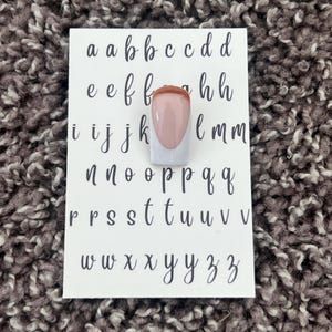 Könnte beinhalten: Eine weiße Karte mit dem Alphabet in schwarzer Schreibschrift. Ein einzelner Nagel mit weißer Basis, nackter Mitte und brauner Spitze befindet sich auf der Karte. Der Hintergrund ist eine strukturierte, graue Oberfläche.