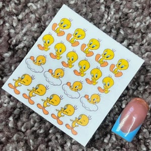 Puede incluir: Una hoja de pegatinas de arte de uñas con el personaje de dibujos animados Tweety Bird en varias poses. Las pegatinas son amarillas con pies y picos naranjas. También se ve una uña pintada con un diseño rosa y azul.