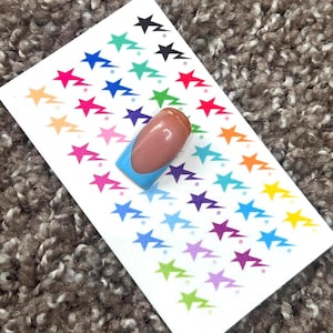 May include: A sheet of nail art stickers featuring colorful star and lightning bolt designs. A single nail with a light blue base and a French tip is placed on the sheet. The nail has a natural-looking pink color.