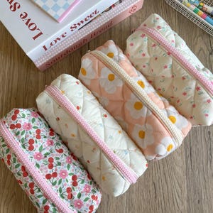 May include: Four quilted pencil cases with floral and cherry patterns. Each case has a pink zipper and a quilted design. The cases are in various colors, including white, pink, and a cherry blossom print. A book with the words "Love Sweet" is in the background.