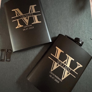 May include: Two black flasks with gold lettering. One flask reads "MICHAEL" above "GROOM" and the date "06.07.2025". The other flask reads "WILLIAM" above "GROOMSMAN" and the date "06.07.2025".