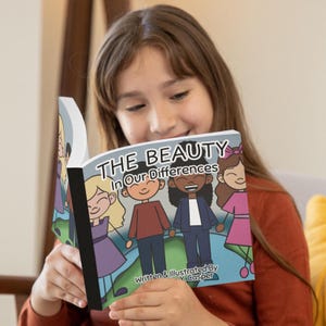 May include: A young girl holds open a children's book titled "THE BEAUTY In Our Differences." The book's cover features cartoon illustrations of diverse children. The text "Written & Illustrated By V. Casper" is also visible.