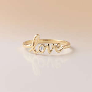 May include: A gold-colored ring spelling out the word "love" in cursive script. The "o" in the word "love" features a small, round, clear gemstone. The ring is set against a soft, neutral background.