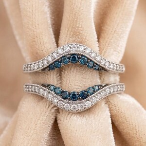 May include: Two curved, silver-toned rings with a wave design. Each ring features a row of small, white gemstones along the outer edges and a row of blue gemstones in the center. The rings are set against a soft, beige background.