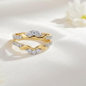 May include: Two-piece gold-tone ring set with a wavy design. The rings are adorned with small, sparkling clear stones. The rings are displayed on a white, silky surface, with white flowers in the background.