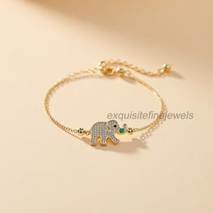 May include: A gold-toned bracelet featuring a small elephant charm. The elephant is encrusted with small, clear stones and has a green accent for the trunk. The bracelet has a delicate chain and adjustable clasp.