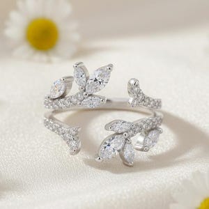 May include: A silver ring with a leaf design, adorned with clear, sparkling gemstones. The ring features marquise-cut stones and smaller round stones, set against a white background with daisy flowers.