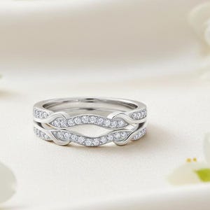May include: Two silver rings with a wave-like design, encrusted with small, sparkling diamonds. The rings are set against a soft, white background, with white flowers in the corners. The rings are a symbol of commitment.