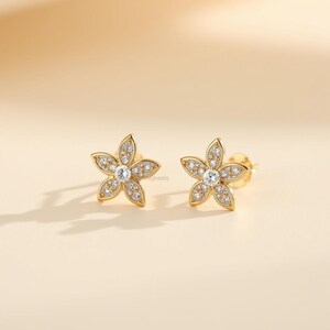 May include: A pair of gold-colored flower-shaped stud earrings. Each earring features five petals encrusted with small, clear gemstones, surrounding a larger, central gemstone. The earrings are set against a soft, neutral background.