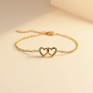 May include: A gold-colored bracelet featuring two intertwined heart-shaped pendants. Each heart is outlined with small, sparkling stones. The bracelet has a delicate chain and a clasp for secure wear, perfect for a romantic gift.