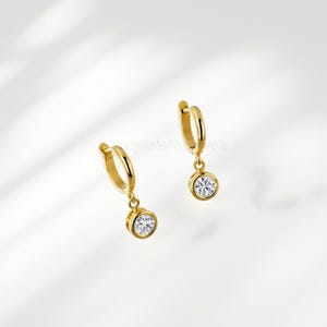 May include: A pair of gold hoop earrings with a dangling, round, clear gemstone. The earrings are set against a soft, white background. The earrings are a classic jewelry design.