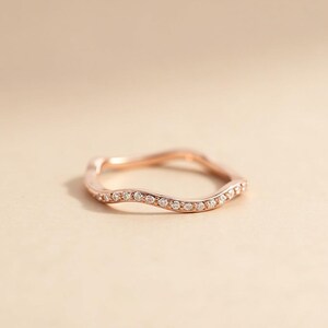 May include: A rose gold ring with a wavy design, set with small, clear gemstones. The ring is displayed on a neutral background, highlighting its delicate and elegant features. The gemstones are evenly spaced along the ring's surface.
