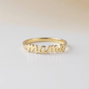 May include: A gold-colored ring with the word "mama" in cursive script. A small diamond is set on the ring's band. The ring is displayed on a white surface with a soft, neutral background. This ring is a piece of jewelry.