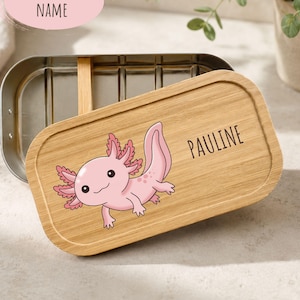 Könnte beinhalten: Eine rechteckige Edelstahl-Lunchbox mit abnehmbarem Bambusdeckel. Der Deckel zeigt eine aufgedruckte Illustration eines rosa Axolotls und den Namen PAULINE in schwarzer Schrift. Der Behälter hat einen Metallverschluss und einen Holztrenner.