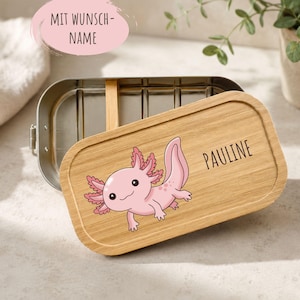 Könnte beinhalten: Eine rechteckige Edelstahl-Lunchbox mit abnehmbarem Bambusdeckel. Der Deckel zeigt eine aufgedruckte Illustration eines rosa Axolotls und den Namen PAULINE. Eine rosa ovale Grafik über der Box enthält den Text MIT WUNSCH-NAME.