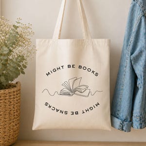 Könnte beinhalten: Eine cremefarbene Stofftasche aus Segeltuch mit zwei Schulterriemen. Auf der Vorderseite befindet sich eine schwarze Strichzeichnung eines offenen Buches, zentriert zwischen dem Text "MIGHT BE BOOKS" oben und "MIGHT BE SNACKS" unten.