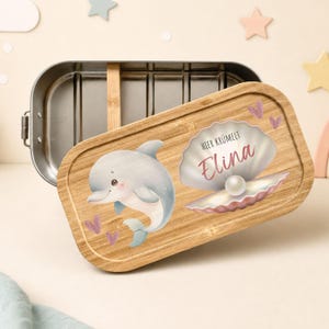 Könnte beinhalten: Eine rechteckige Edelstahl-Lunchbox mit einem Bambusdeckel. Der Deckel zeigt eine aufgedruckte Illustration eines grauen Delfins, einer Muschel mit einer Perle und den Text "HIER KRÜMELT Elina" in Rosa und Schwarz, verziert mit kleinen Herzen.