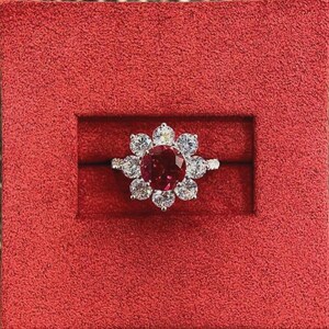 May include: A flower-shaped ring with a large, round, deep red gemstone at the center, surrounded by smaller, clear gemstones. The ring is set in a silver band and presented in a red velvet box.