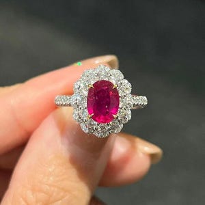 Puede incluir: Primer plano de un anillo con una gran gema de rubí ovalada, rodeada por un halo de pequeños diamantes brillantes. La banda del anillo también está adornada con diamantes, creando una apariencia lujosa y elegante. El anillo está engastado en oro o platino.