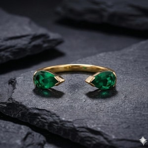 May include: A gold ring featuring two pear-shaped, vibrant green gemstones. The ring has an open design, with the gemstones set at opposite ends of the band. The ring is displayed on a dark, textured surface.