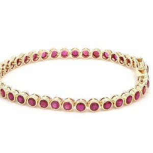 Puede incluir: Una pulsera dorada con una serie de piedras preciosas rojas redondas. Las piedras están engastadas juntas, creando una línea continua alrededor de la muñeca. La pulsera tiene un cierre seguro.