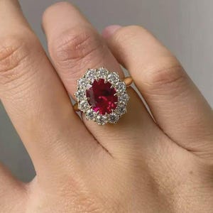 May include: A gold ring featuring a large, oval-cut, deep red gemstone surrounded by a halo of smaller, clear gemstones. The ring is set on a gold band, creating a classic and elegant design. The ring is shown on a person's finger.