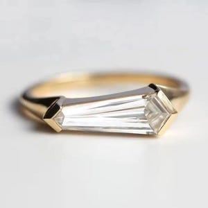 May include: A gold ring with a clear, rectangular-cut gemstone. The gemstone is set horizontally, with a gold band and geometric accents on each end. The ring is displayed on a white background.