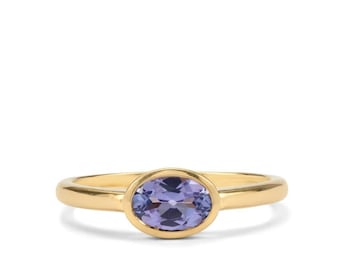 Puede incluir: Un anillo de oro con una banda lisa y pulida, que presenta una piedra preciosa ovalada y facetada de color púrpura engastada en un bisel. El anillo tiene un diseño sencillo con un cuerpo redondeado y una piedra púrpura brillante.