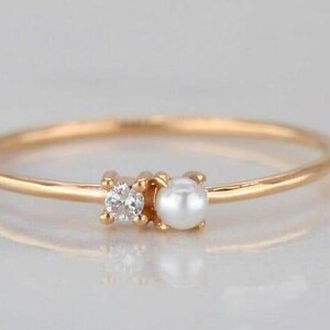 May include: A delicate gold ring featuring a small diamond and a white pearl. The ring has a thin band and is set against a soft, neutral background. This elegant piece of jewelry is perfect for everyday wear.