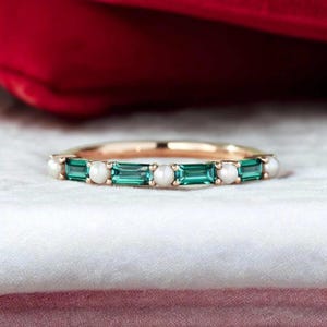 Könnte beinhalten: Ein goldener Bandring mit abwechselnd smaragdgrünen Edelsteinen und kleinen weißen Perlen. Der Ring ist vor einem weißen und roten Hintergrund platziert, der das Design und die Farbe des Schmucks hervorhebt.