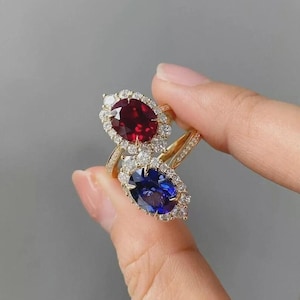 May include: Two gold rings with large, oval-cut gemstones. One ring features a deep red gemstone, the other a vibrant blue. Both are surrounded by smaller, clear stones, creating a halo effect. The rings are held against a gray background.