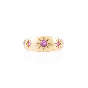May include: A gold-colored ring featuring three starburst designs. Each starburst has a central pink gemstone. The ring has a smooth, polished surface and a rounded shape. The ring is likely made of gold and is a piece of jewelry.