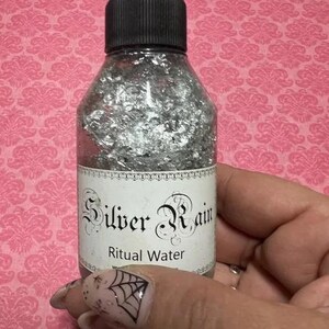 以下が含まれることがあります： 銀色のフレークが入った透明なガラス瓶と黒いキャップ。ラベルには「Silver Rain Ritual Water」と書かれています。背景はピンク色の花柄です。