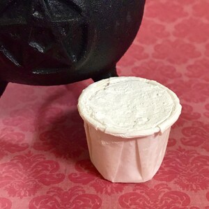 Op de afbeelding: Een kleine, witte papieren beker gevuld met een witte substantie, mogelijk een poeder of pasta. De beker heeft een geplooid ontwerp en staat op een roze oppervlak met patronen. Een zwarte ketel is gedeeltelijk zichtbaar op de achtergrond.