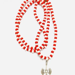 May include: A necklace featuring alternating red and white beads. The necklace has a silver-colored pendant in the shape of a double-bladed axe. The beads are small and round, creating a striking contrast with the silver pendant.