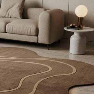 May include: A modern living room scene featuring a beige sofa, a marble side table with a gold lamp, and a brown rug with a unique, flowing design. The rug has light beige wavy lines.