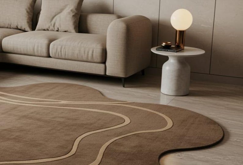 May include: A modern living room scene featuring a beige sofa, a marble side table with a gold lamp, and a brown rug with a unique, flowing design. The rug has light beige wavy lines.