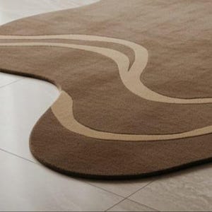 May include: A brown area rug with a unique, organic shape. The rug features a lighter beige, flowing design across its surface. The rug is placed on a light-colored tile floor, adding a touch of modern design to any space.