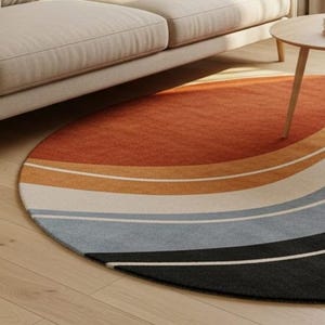 May include: A round area rug with a retro design. The rug features a gradient of colors, starting with a burnt orange at the top and transitioning through shades of tan, white, light blue, gray, and black at the bottom. The rug is placed on a light wood floor.