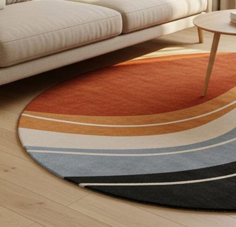 May include: A round area rug with a retro design. The rug features a gradient of colors, starting with a burnt orange at the top and transitioning through shades of tan, white, light blue, gray, and black at the bottom. The rug is placed on a light wood floor.