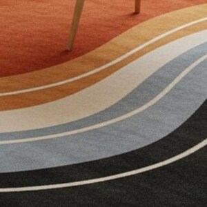 May include: A colorful area rug with a wavy design. The rug features bands of orange, brown, tan, white, gray, and black. A wooden table leg is visible in the upper portion of the image.
