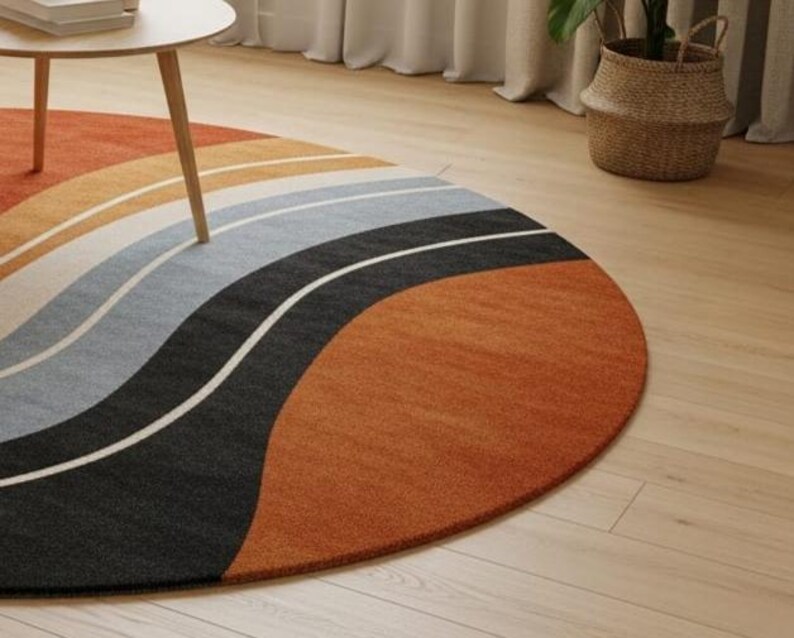 May include: A round area rug with a wave design in shades of orange, tan, gray, black, and white. The rug is placed on a light wood floor, with a small table and a woven basket in the background.