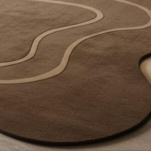 May include: A brown, uniquely shaped rug with a wavy, lighter-colored design. The rug has a dark brown border and a soft, textured surface. The design is reminiscent of a flowing river or abstract landscape, adding a modern touch to any room.