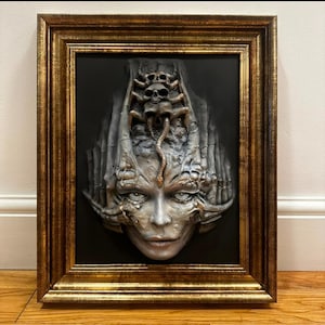 Puede incluir: Una escultura de pared enmarcada que presenta un rostro humano central con protuberancias orgánicas texturizadas y una pequeña figura similar a una serpiente que se extiende desde una corona con calaveras, dentro de un marco dorado ornamentado.