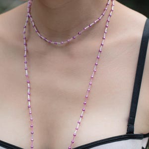 May include: A long necklace featuring a delicate chain of small, translucent pink beads interspersed with metallic purple spheres. The necklace ends in two symmetrical drops containing faceted purple stones, gold-toned accents, and small white pearls.