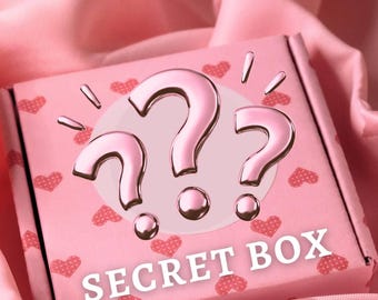 May include: A square pink cardboard box featuring a repeating pattern of small red hearts. The center of the box displays three large, metallic pink question marks above the white text "SECRET BOX" printed in a bold, sans-serif font.