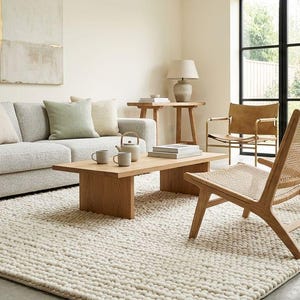 Puede incluir: Salón de tonos neutros con un sofá gris claro, una mesa de centro de madera y dos sillas tejidas. Una gran alfombra blanquecina cubre el suelo. Cojines en tonos neutros y verdes adornan el sofá. Una planta en maceta añade un toque de vegetación.