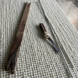 Puede incluir: Una herramienta larga y plana de madera marr&oacute;n con una muesca en forma de V en ambos extremos, un peque&ntilde;o mango de madera marr&oacute;n oscuro con una punta de metal curvada envuelta en cuerda clara, y dos varillas met&aacute;licas plateadas largas y finas.