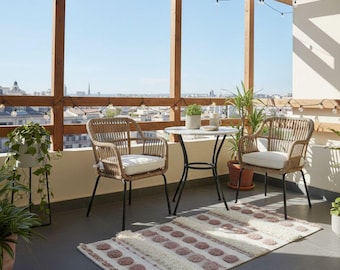 Puede incluir: Un conjunto de balcón con dos sillas de mimbre, una pequeña mesa redonda y una alfombra estampada. Las sillas tienen cojines blancos. El balcón está rodeado por una estructura de madera con luces de cadena y plantas en macetas. El fondo muestra un paisaje urbano.